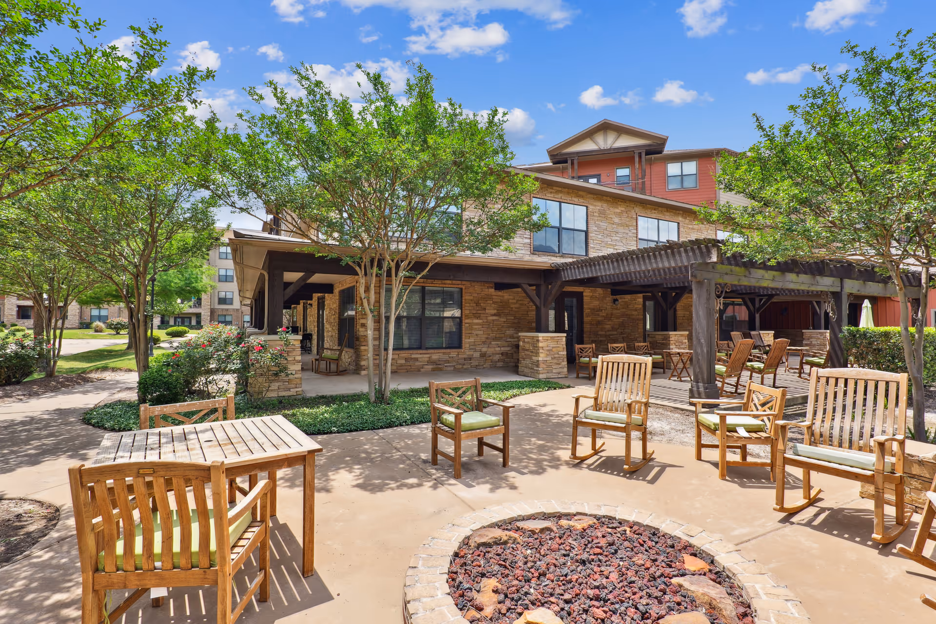 Outdoor seating area at a senior living facility with wooden chairs and tables arranged around a circular fire pit. The area is shaded by trees and a pergola attached to a stone and wood building under a blue sky with scattered clouds.