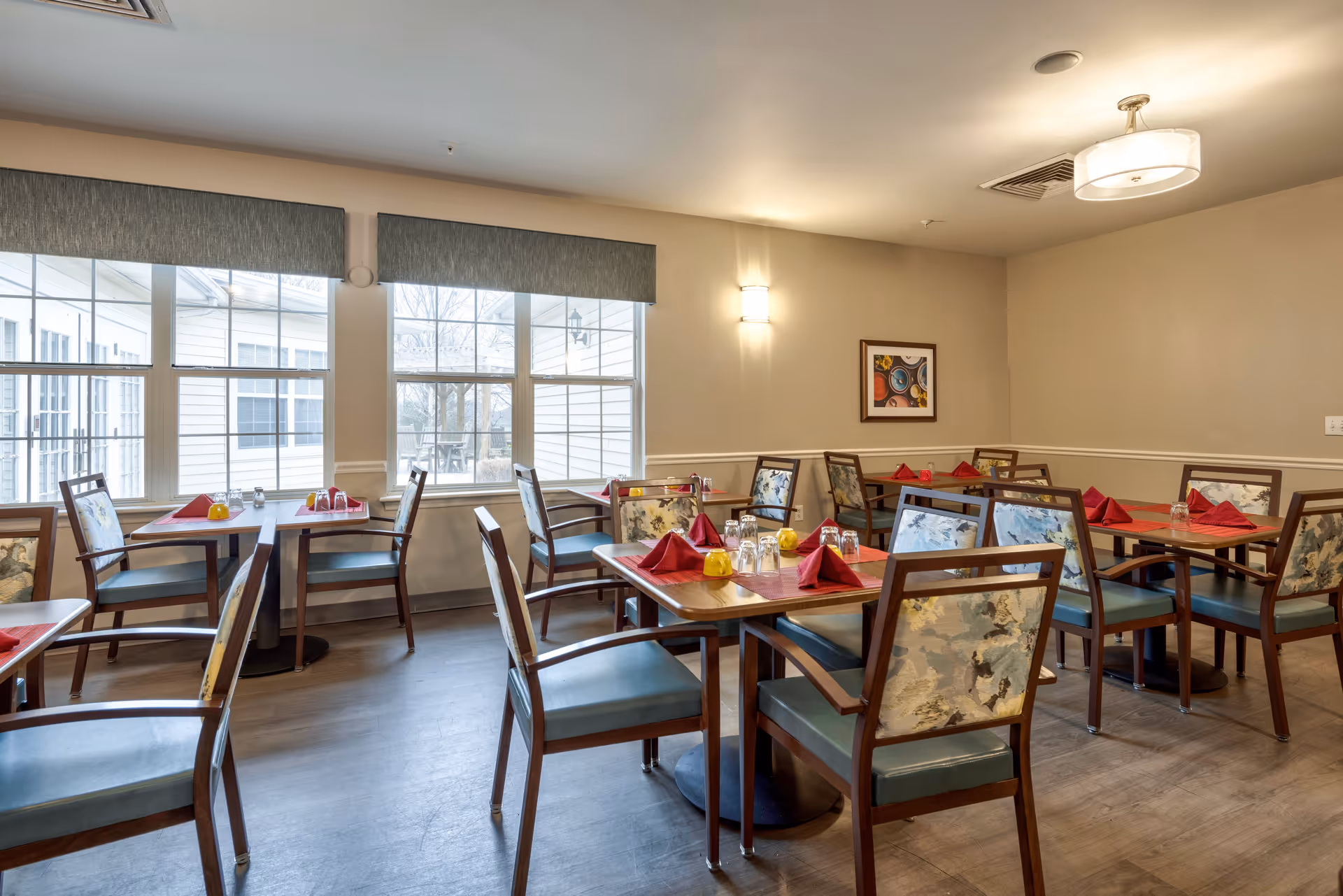 A dining room with several wooden tables and chairs arranged neatly. Each table is set with red folded napkins, glasses, and small yellow condiment containers. Large windows on one side allow natural light to fill the room. The walls are painted beige with a framed artwork hanging on one wall and ceiling lights providing additional illumination.