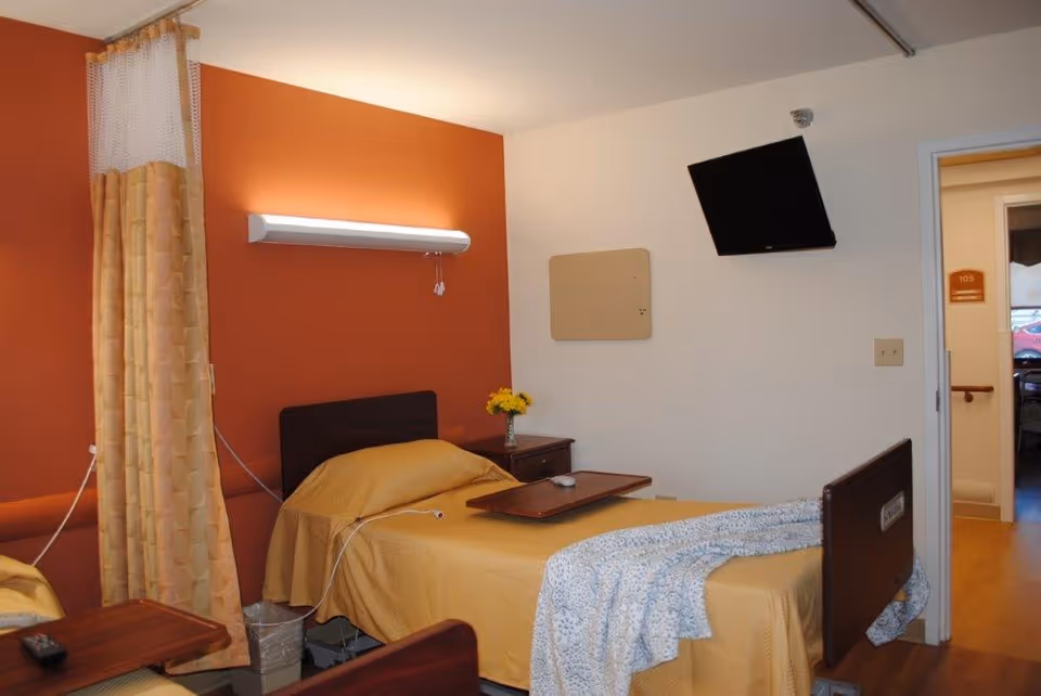 A senior living facility bedroom with a single bed covered in a yellow blanket and a blue patterned throw. There is a wooden bedside table with a vase of yellow flowers, a wall-mounted TV, a light fixture above the bed, and a privacy curtain partially drawn. The room has an orange accent wall and a door leading to a hallway.
