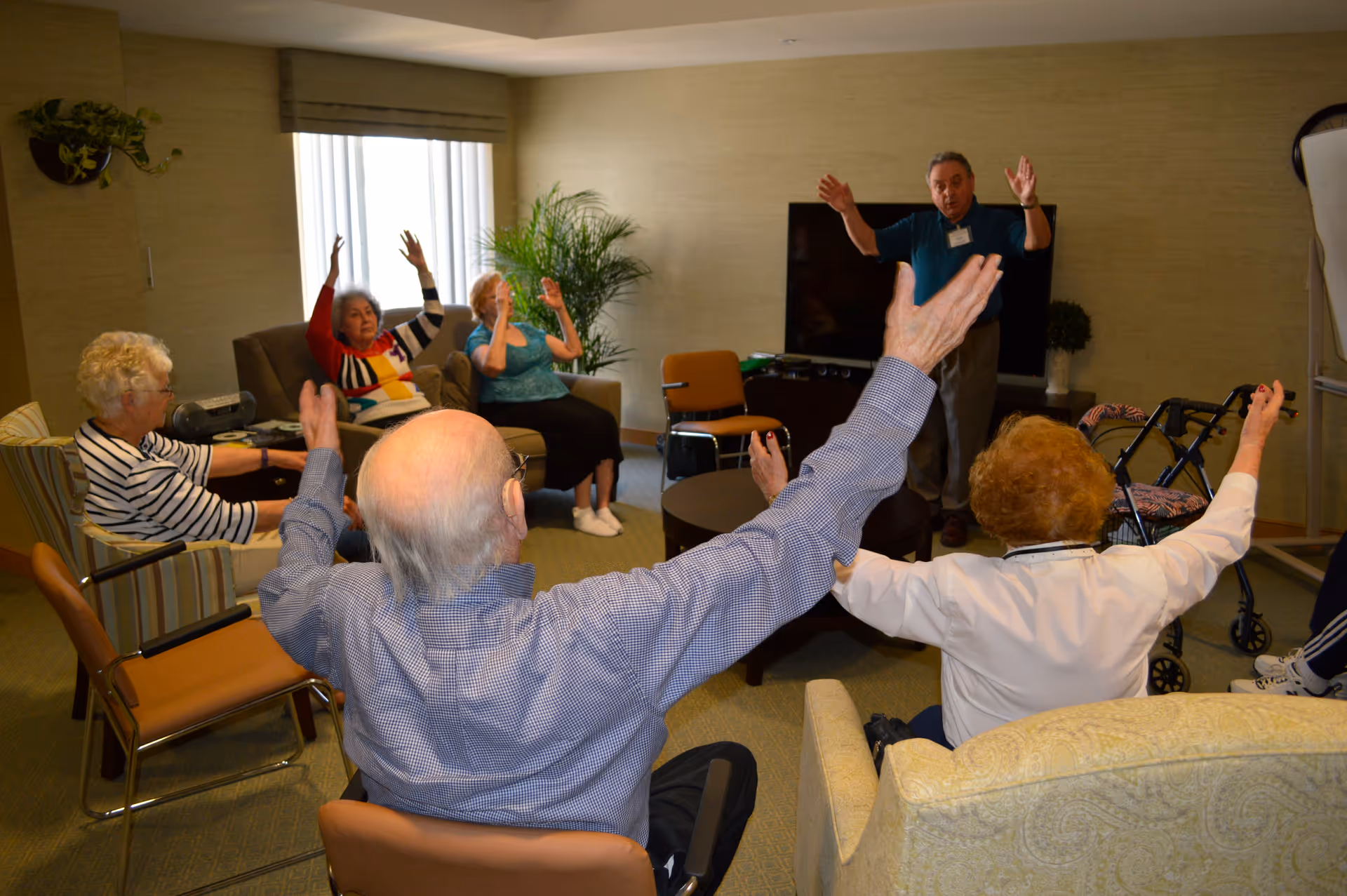 A group of elderly people seated in a living room area participating in a seated exercise or stretching session led by an instructor standing near a television. The participants have their arms raised and appear engaged in the activity.