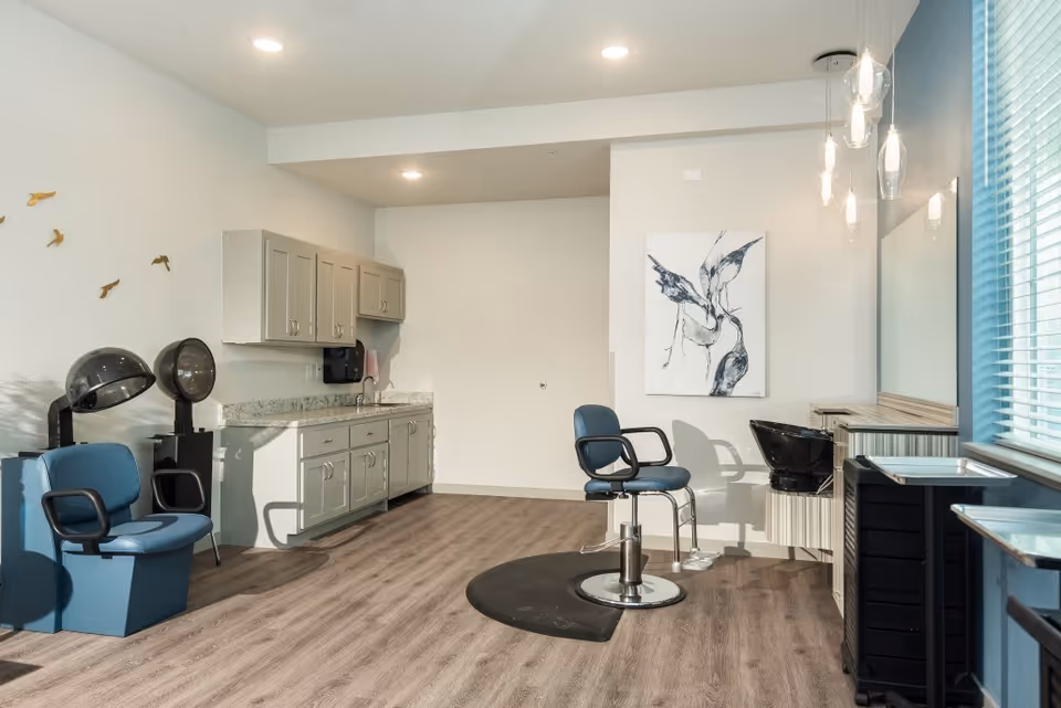 Interior of a salon area in a senior living facility with two blue salon chairs, two hair dryers, a countertop with cabinets, a black wash basin, a large mirror, and modern pendant lights. There is a window with blinds on the right side and a piece of abstract art on the wall.