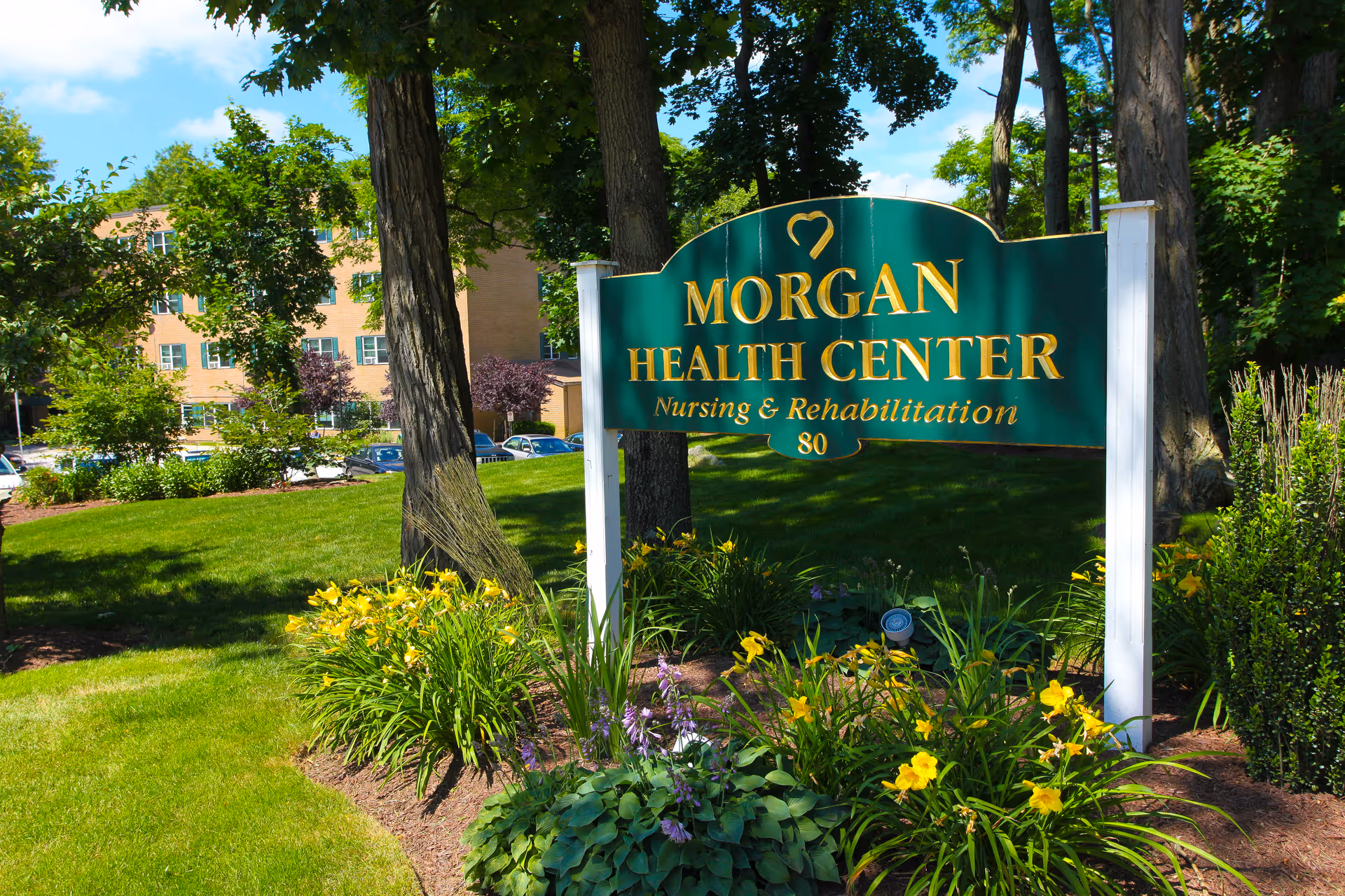 Outdoor view of a green and gold sign for Morgan Health Center Nursing & Rehabilitation surrounded by trees, grass, and flowering plants with a building and parked cars in the background.