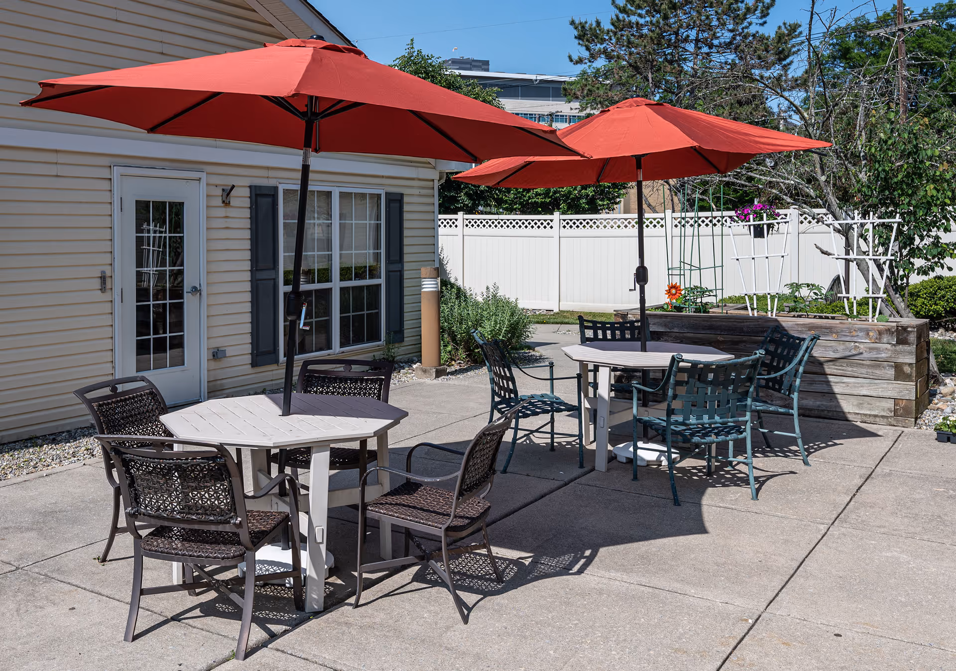 Outdoor patio area with two round tables, each shaded by a large red umbrella. Each table is surrounded by several chairs. The patio is adjacent to a building with beige siding, a white door, and a window with dark shutters. There is a white fence and some greenery in the background.