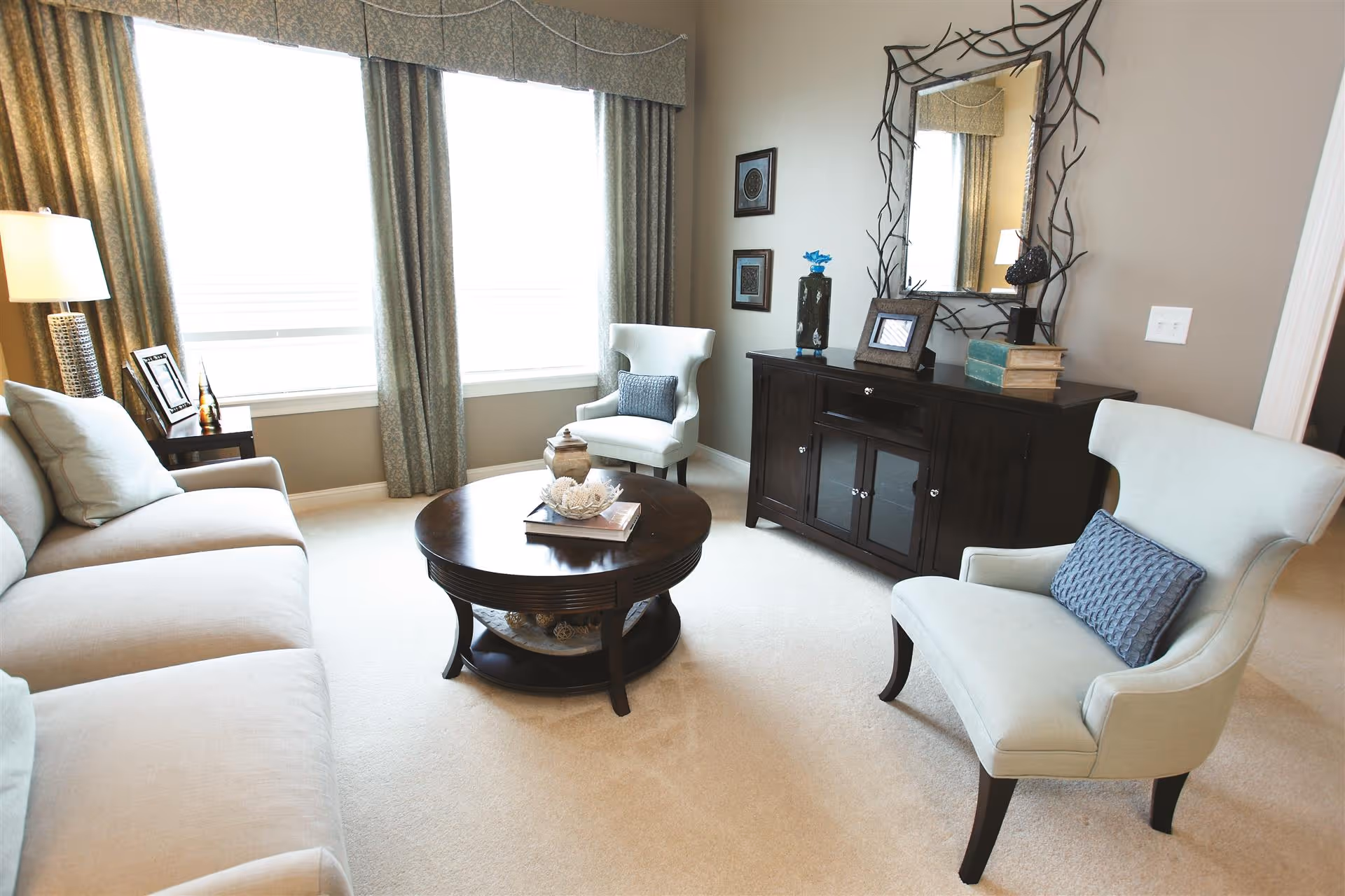 A bright and cozy living room with a beige sofa, two light blue armchairs with blue cushions, a round dark wooden coffee table with decorative items, a dark wooden cabinet with books and framed pictures, and a large decorative mirror on the wall. Large windows with curtains allow natural light to fill the room.