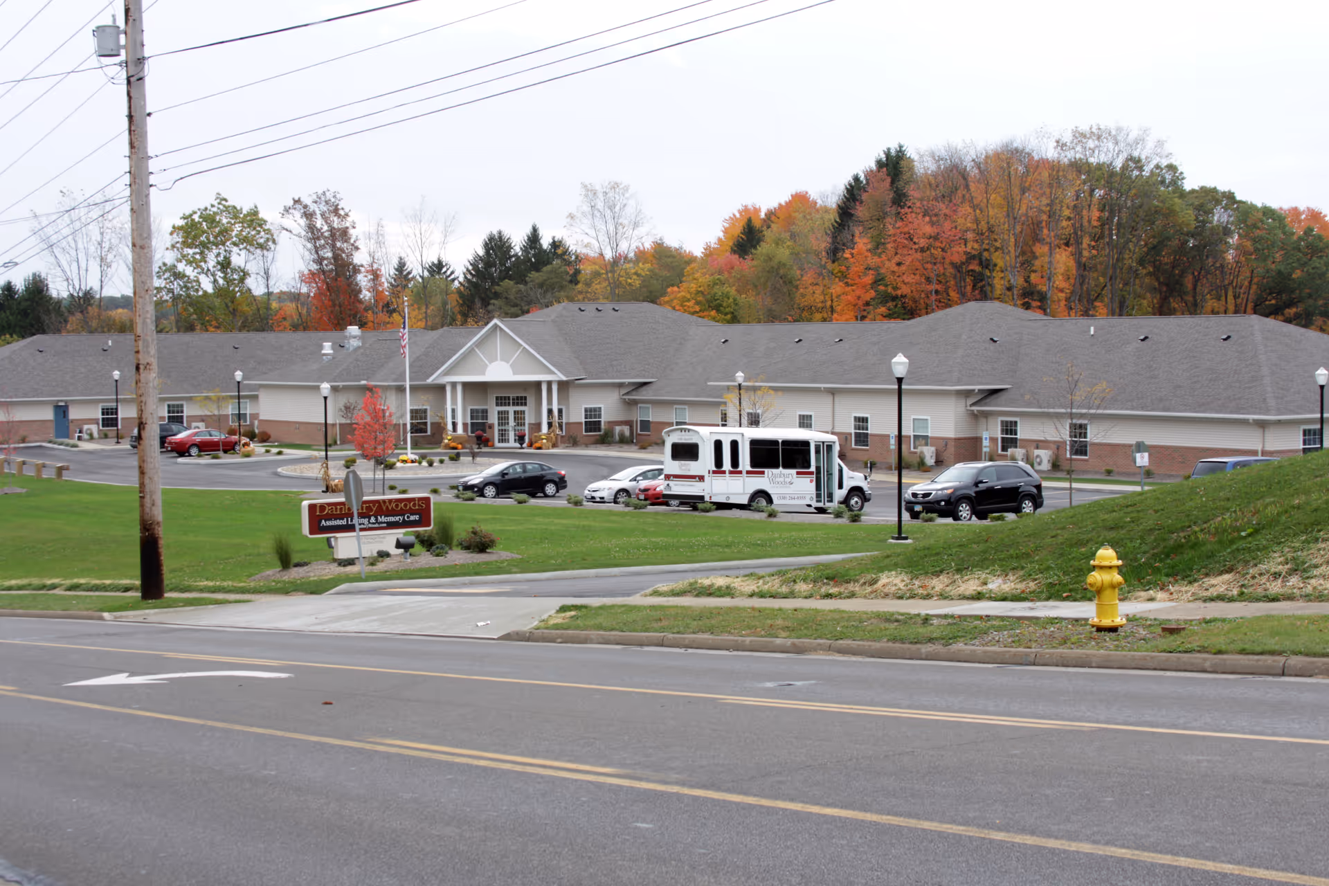 Exterior view of Danbury Woods Assisted Living & Memory Care facility with a parking lot containing several cars and a shuttle bus. The building is a single-story structure with a gray roof and beige walls. Trees with autumn foliage are visible in the background, and a yellow fire hydrant is on the grassy area near the road in the foreground.