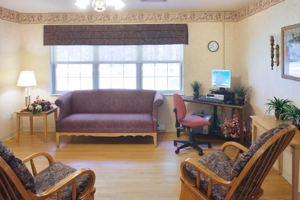 A cozy living room with a purple upholstered sofa against a large window with blinds. Two wooden armchairs with floral cushions face the sofa. A wooden side table with a lamp and flower arrangement is next to the sofa. A computer desk with a desktop computer, keyboard, and a red office chair is positioned against the wall. The room has light-colored walls with a decorative border near the ceiling, a wall clock, framed artwork, and several potted plants.