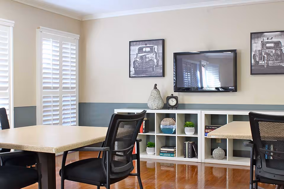 A bright room with two tables and black mesh chairs, a white cubby shelf with decorative items and books, a wall-mounted flat screen TV, and two black and white framed pictures of vintage trucks on the wall. The room has wooden flooring and white plantation shutters on the windows.