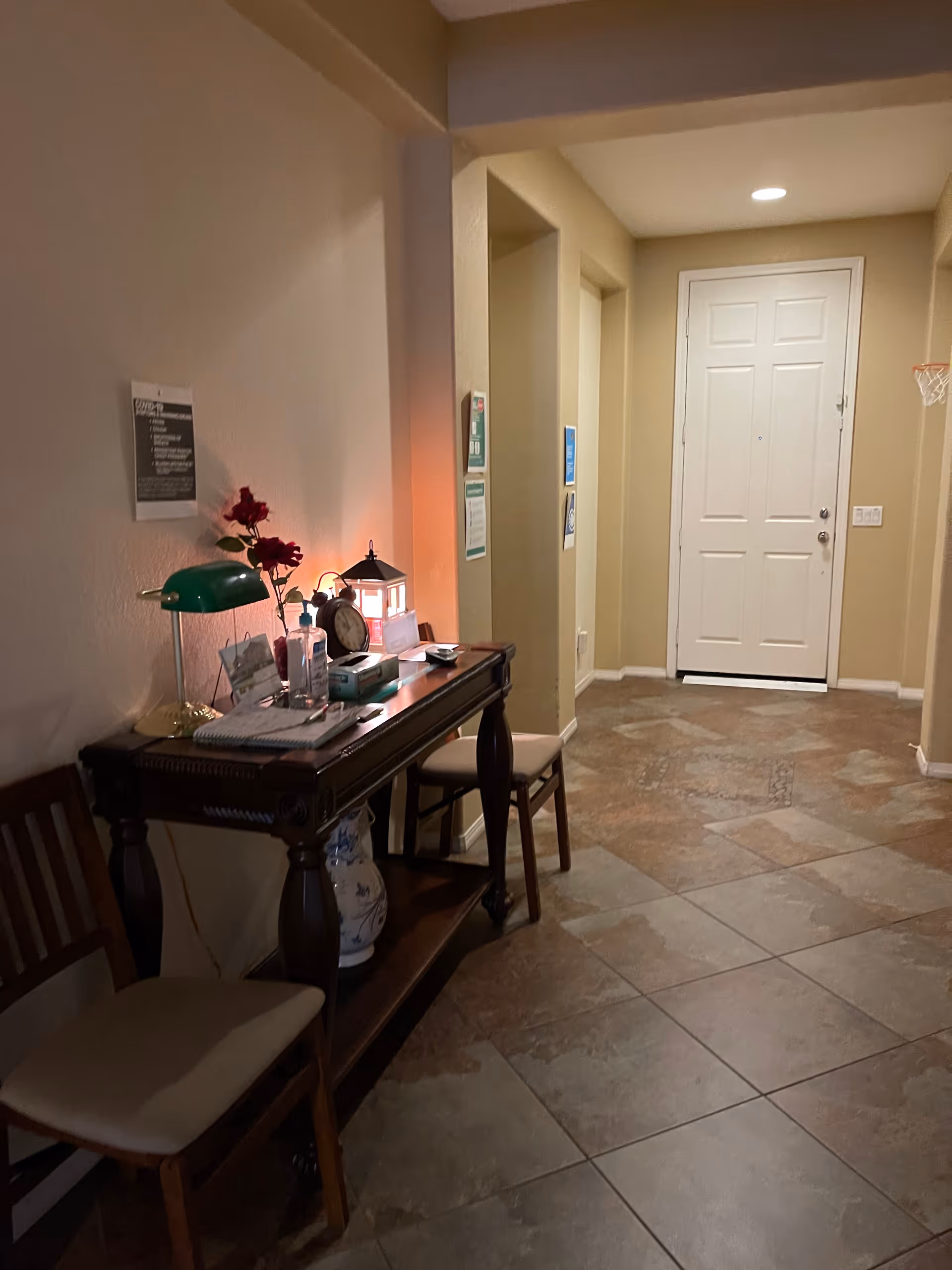 A hallway with a tiled floor leading to a white door at the end. On the left side, there is a dark wooden console table with various items on it including a green desk lamp, a clock, a small lantern, and some papers. Two wooden chairs with cushions are placed next to the table. The walls are painted beige and there are some signs and notices posted on the walls.