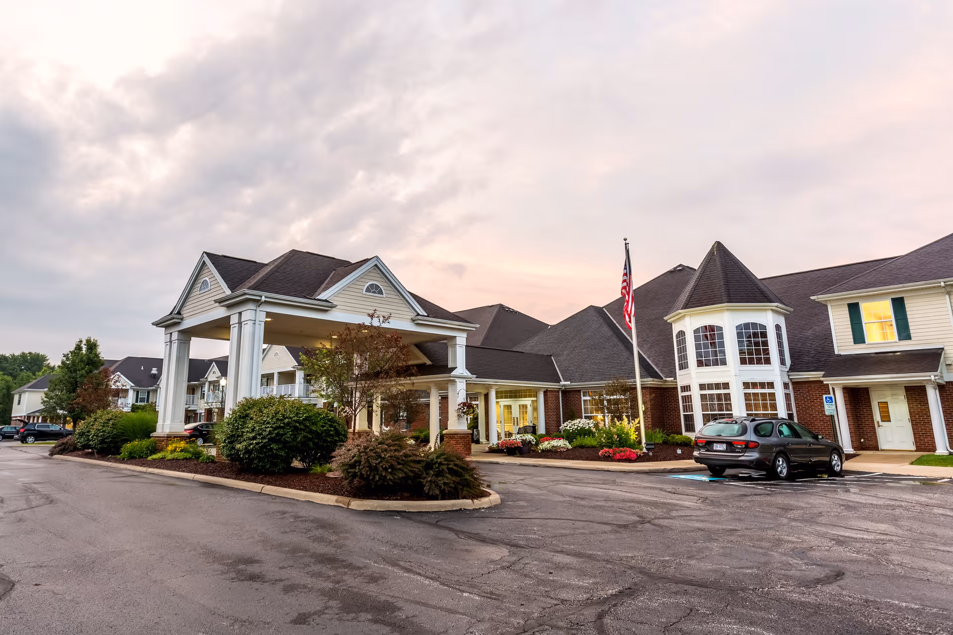 Exterior view of Independence Village of Aurora senior living facility at dusk, showing a large building with a covered entrance, landscaped bushes and flowers, an American flag, and parked cars in front.