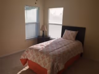 A small bedroom with a single bed covered with a patterned quilt and a pillow. There is a bedside table with a lamp between two windows with blinds partially drawn. The walls are light-colored and the floor is carpeted.