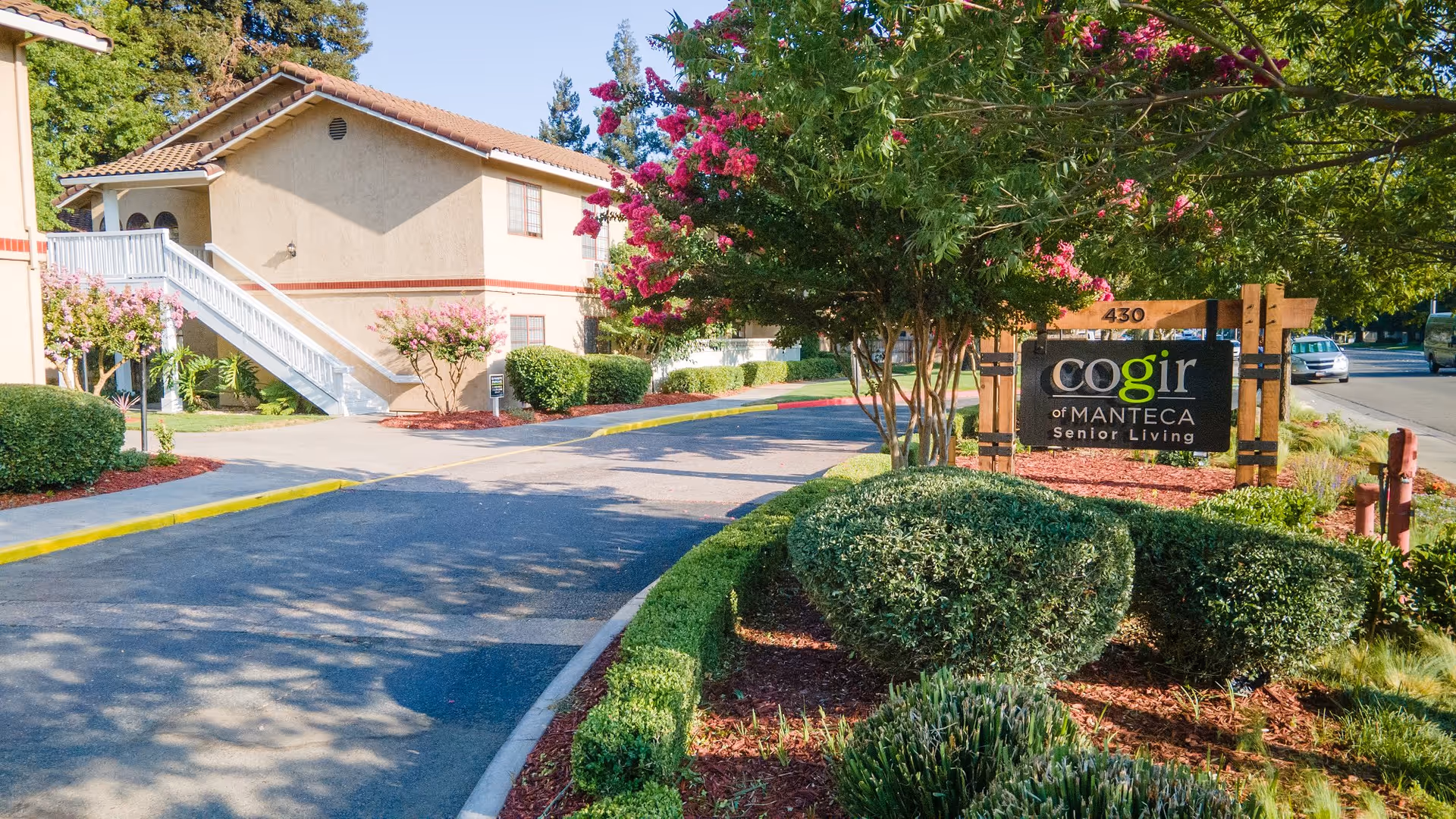 Exterior view of Cogir of Manteca senior living facility showing a driveway lined with manicured bushes and trees, a building with beige walls and a tiled roof, and a sign that reads 'Cogir of Manteca Senior Living'.