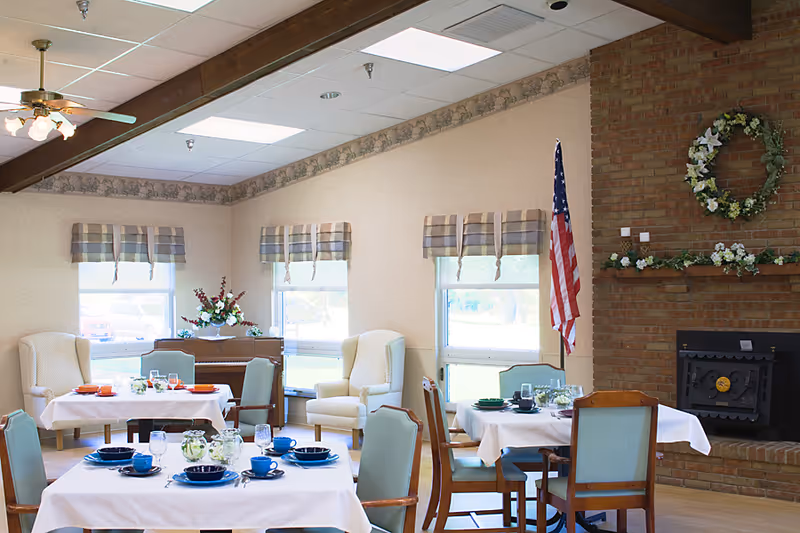 A cozy dining room in a senior living facility with tables set for meals, featuring blue and orange dishware. The room has large windows with plaid valances, two cream-colored armchairs, a brick fireplace decorated with a floral wreath and garland, and an American flag standing beside the fireplace.