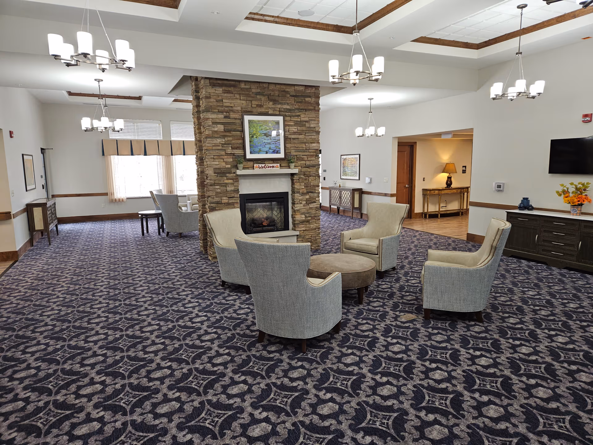 A spacious senior living facility common area with a patterned carpet, four upholstered armchairs arranged around a round ottoman in front of a stone fireplace. The room features multiple chandeliers, large windows with curtains, framed artwork, and a flat-screen TV mounted on the wall. There are wooden cabinets and a table with a lamp in the background.
