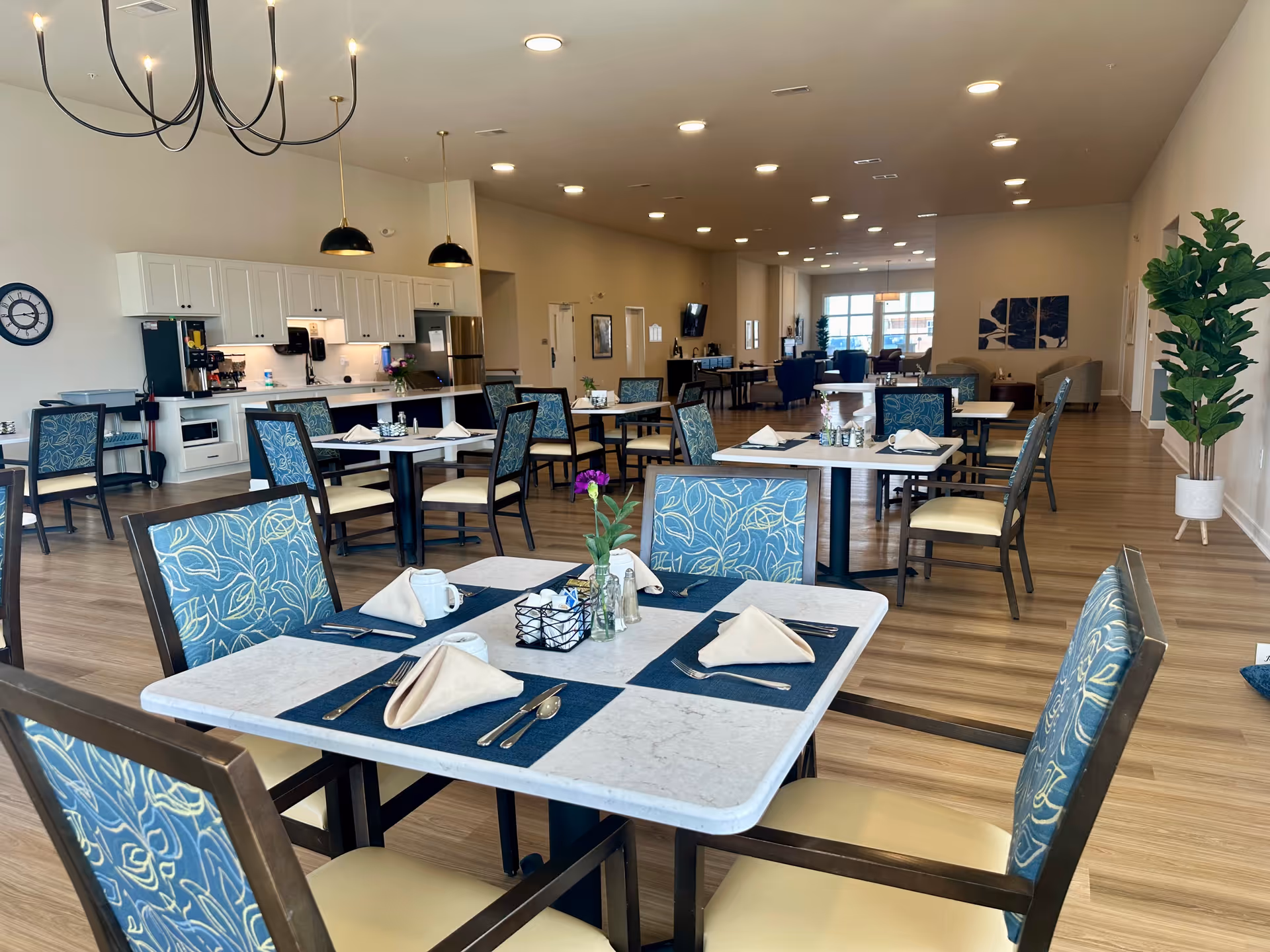A spacious dining room in a senior living facility with multiple tables set with blue placemats, beige napkins, silverware, and small flower vases. The room features wooden flooring, white cabinetry along one wall with kitchen appliances, and modern pendant lights hanging from the ceiling. There are comfortable chairs with blue patterned upholstery and a large plant in a white pot near the wall.