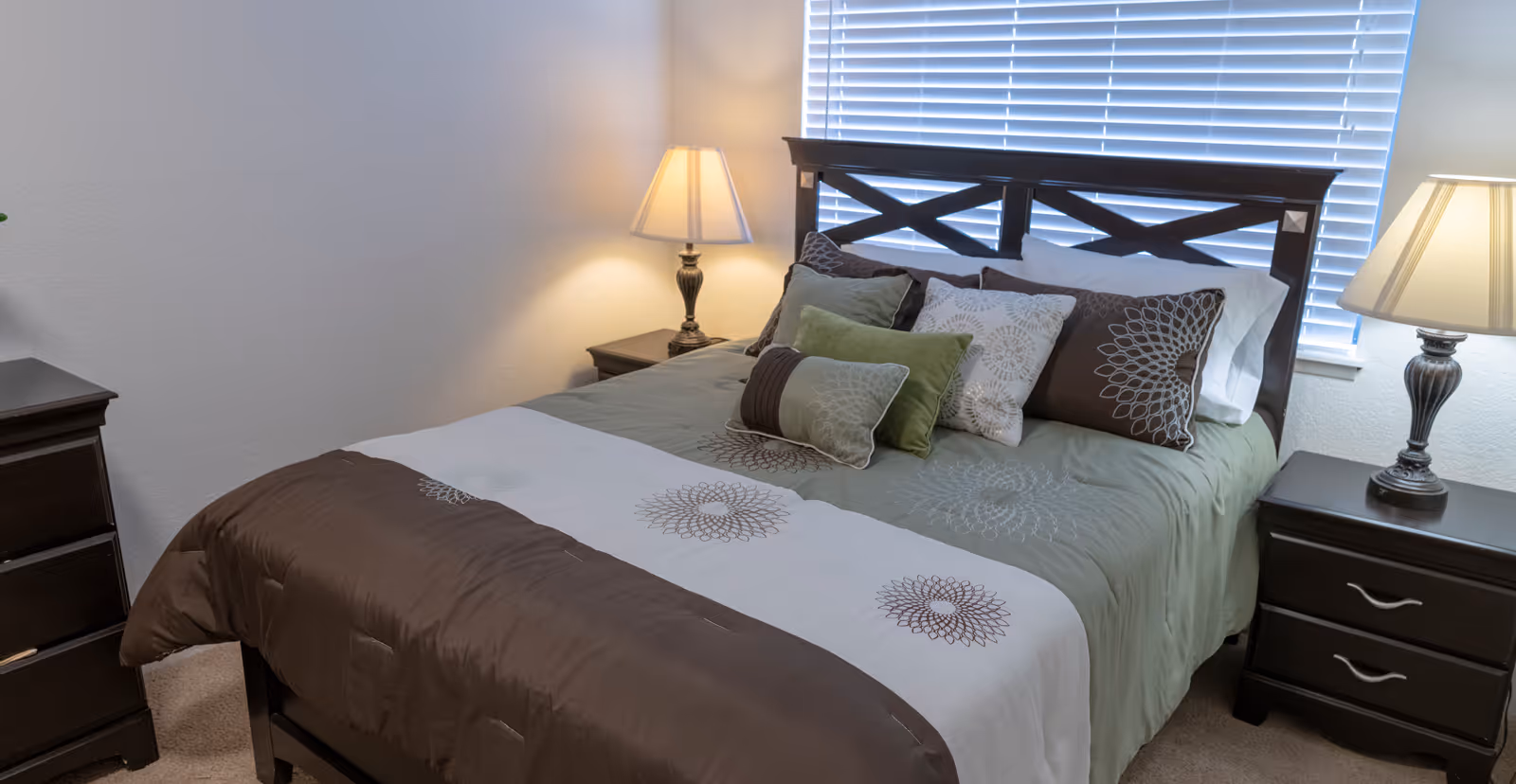 Neatly made bed with decorative pillows flanked by two nightstands and lamps in a bedroom with window blinds.
