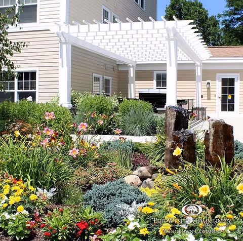 Landscaped garden with colorful flowers, a stone water feature, and a white pergola attached to a beige senior living building.