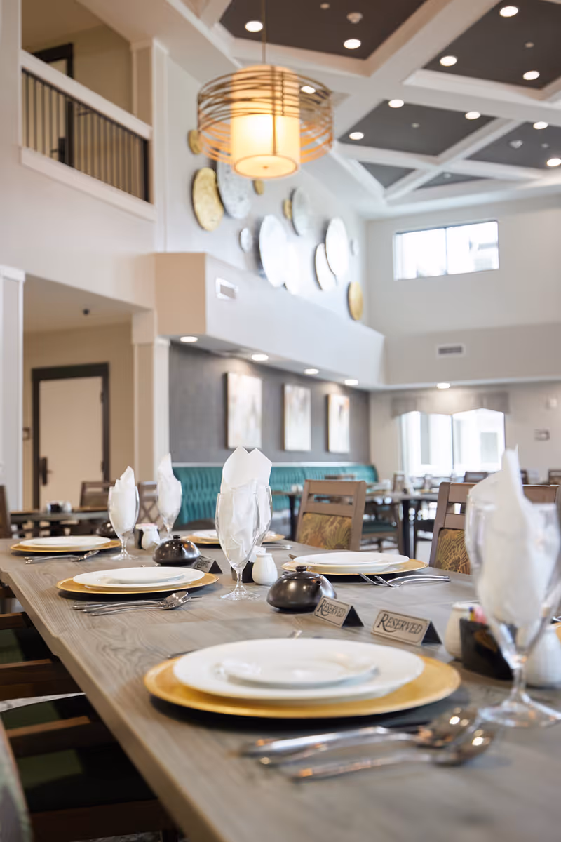 A dining room in a senior living facility with a long wooden table set with plates, gold chargers, silverware, glasses with folded white napkins, and reserved signs. The room features high ceilings with recessed lighting, a modern hanging light fixture, decorative wall art, and large windows letting in natural light.