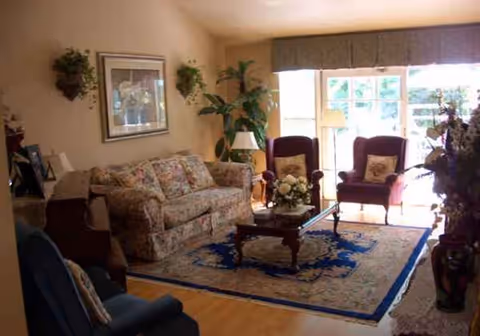 Cozy senior living room with floral sofa, armchairs, coffee table, plants, and a large window.