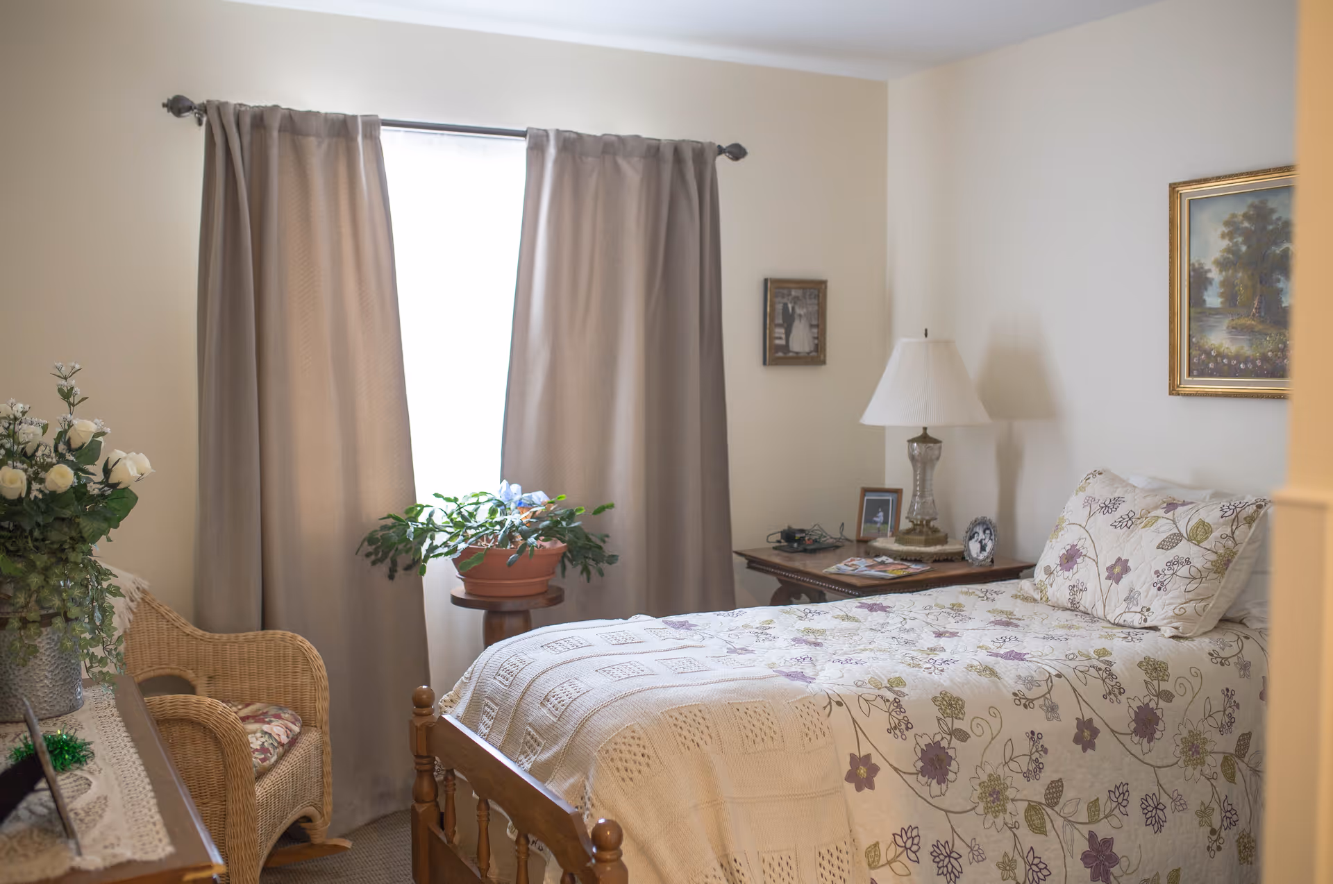 A cozy bedroom with a single wooden bed covered in a floral quilt. There is a wicker chair with a cushion, a small round table with a potted plant, and a wooden nightstand with a lamp, framed photos, and other small items. Beige curtains cover the window, and framed artwork hangs on the walls.