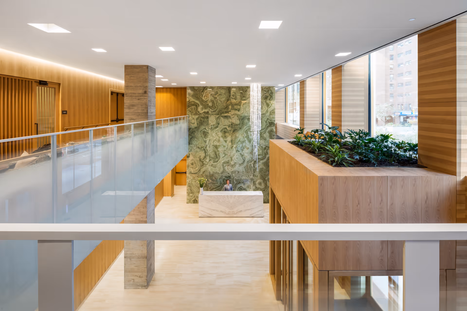 Modern two-story lobby with a marble reception desk, green stone feature wall, glass railings, and planter boxes by large windows.