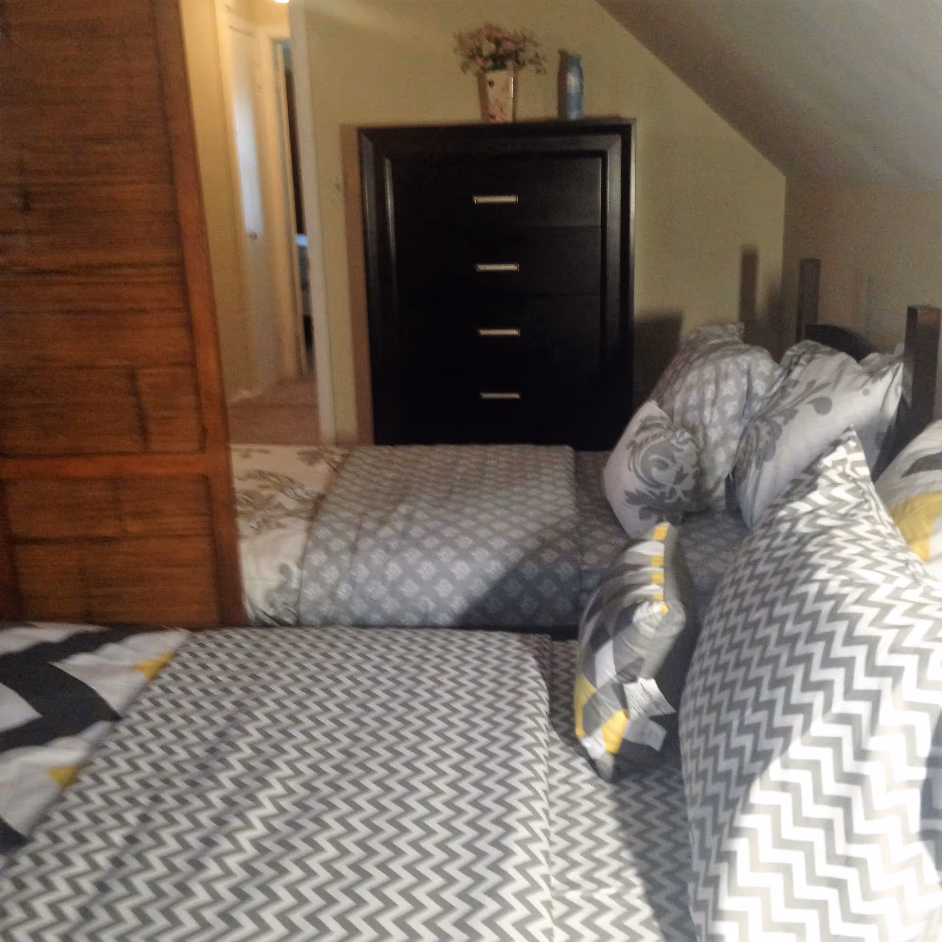 A bedroom with a bed covered in patterned bedding and multiple pillows. There is a dark wooden dresser with four drawers against the wall, topped with a vase of flowers and a bottle. A wooden closet door is partially visible on the left side, and a hallway can be seen in the background.