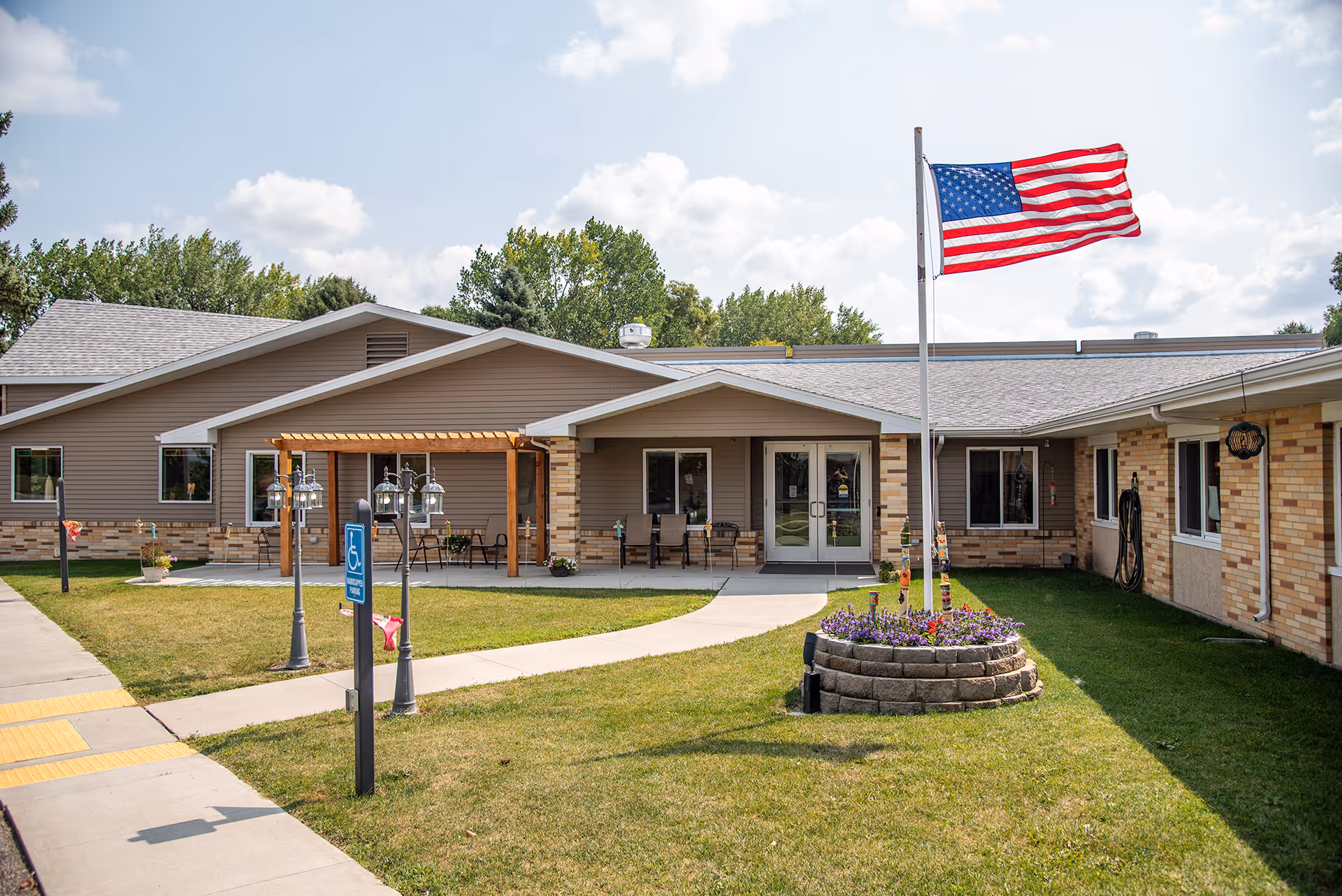Front entrance of a single-story senior living facility with an American flag, a curved walkway, a small flower bed, and a covered seating area.