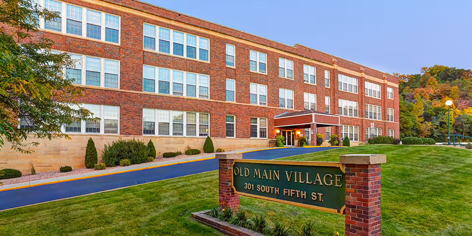 Brick three-story senior living building with a front lawn and a sign reading "Old Main Village 301 South Fifth St."