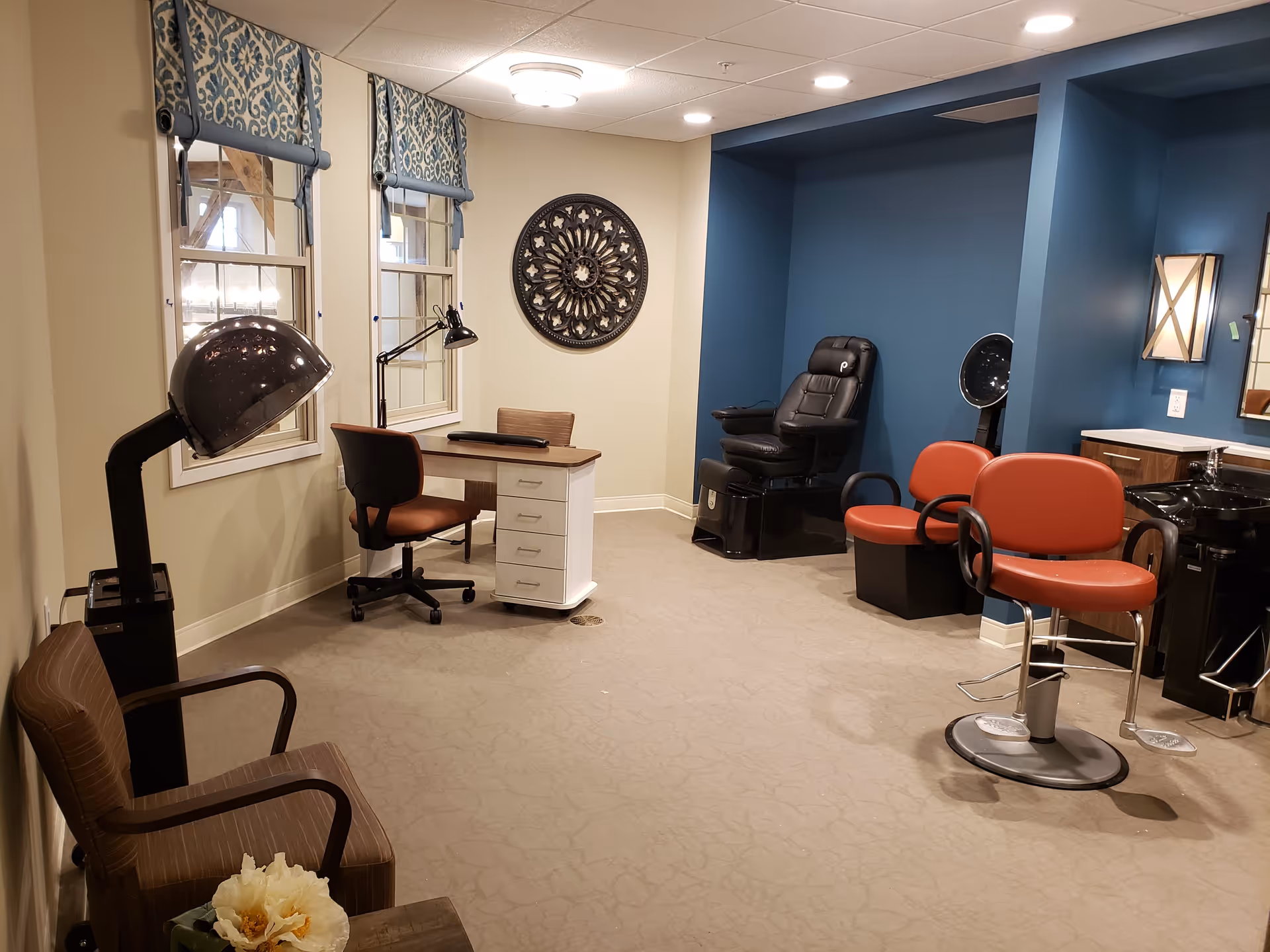 Interior of a senior living facility's salon area with beige walls and blue accent wall. The room features a hair dryer chair, a manicure table with a rolling chair, two red salon chairs, a black shampoo sink, and a decorative wall piece. There are two windows with patterned blue and white valances and a ceiling light fixture.