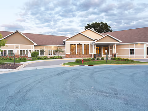 Exterior view of a single-story senior living facility building with beige siding and a brown roof under a cloudy sky. The entrance has large windows and a covered porch area with a driveway and landscaped greenery in front.