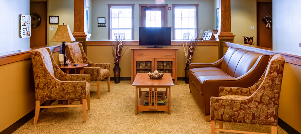 Communal sitting area with upholstered chairs, a leather sofa, coffee table and a television between two windows.