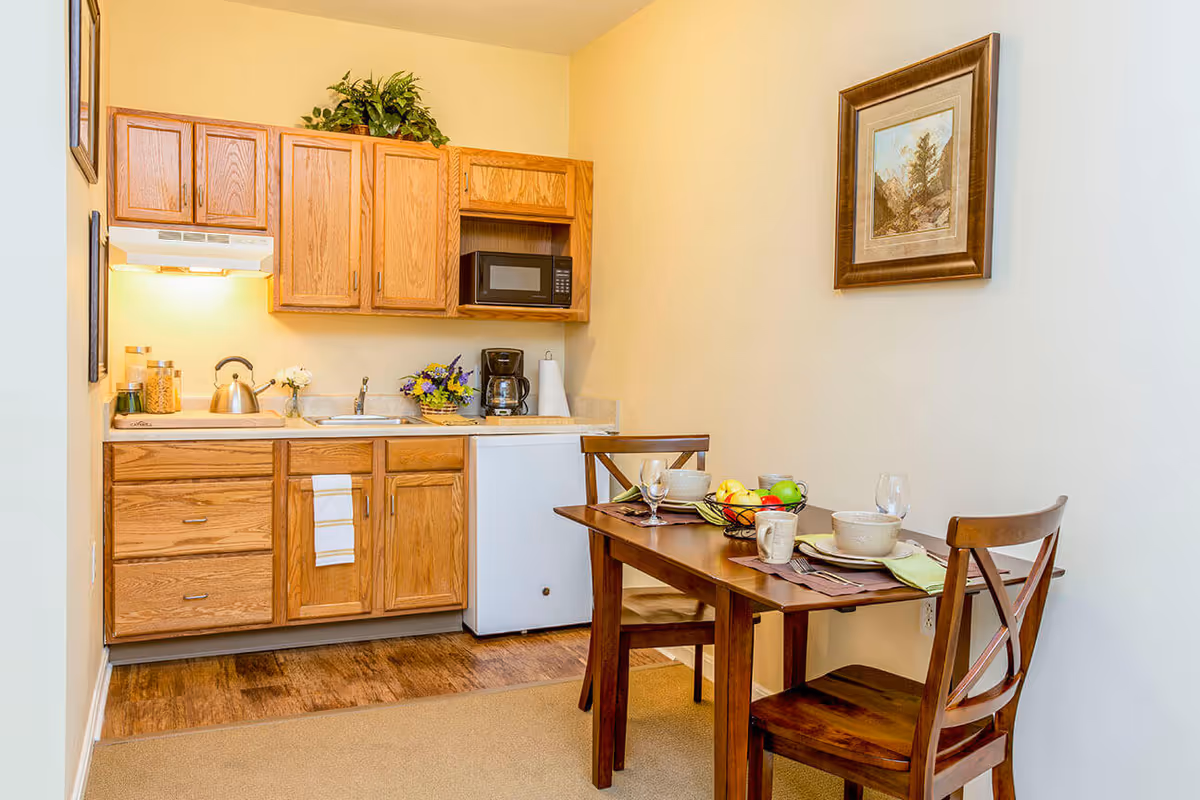 Small kitchenette with oak cabinets, a microwave and mini-fridge, and a wooden dining table set for two.
