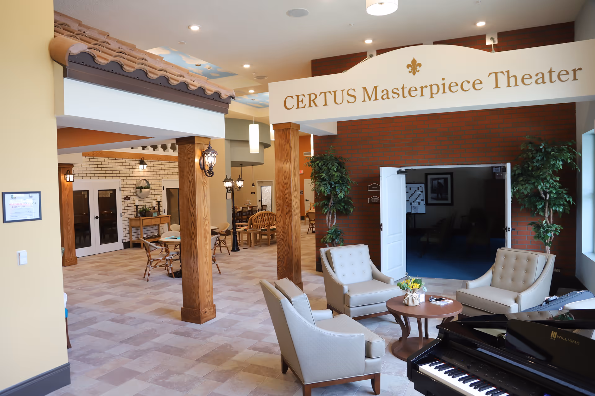 Interior view of CERTUS Premier Memory Care Living featuring a seating area with four beige armchairs around a small round wooden table with a flower arrangement. A black grand piano is visible in the foreground. The space has wooden pillars, tiled flooring, and a sign overhead that reads 'CERTUS Masterpiece Theater' above double doors leading to a theater room. Additional tables and chairs are visible in the background along with decorative lighting and plants.