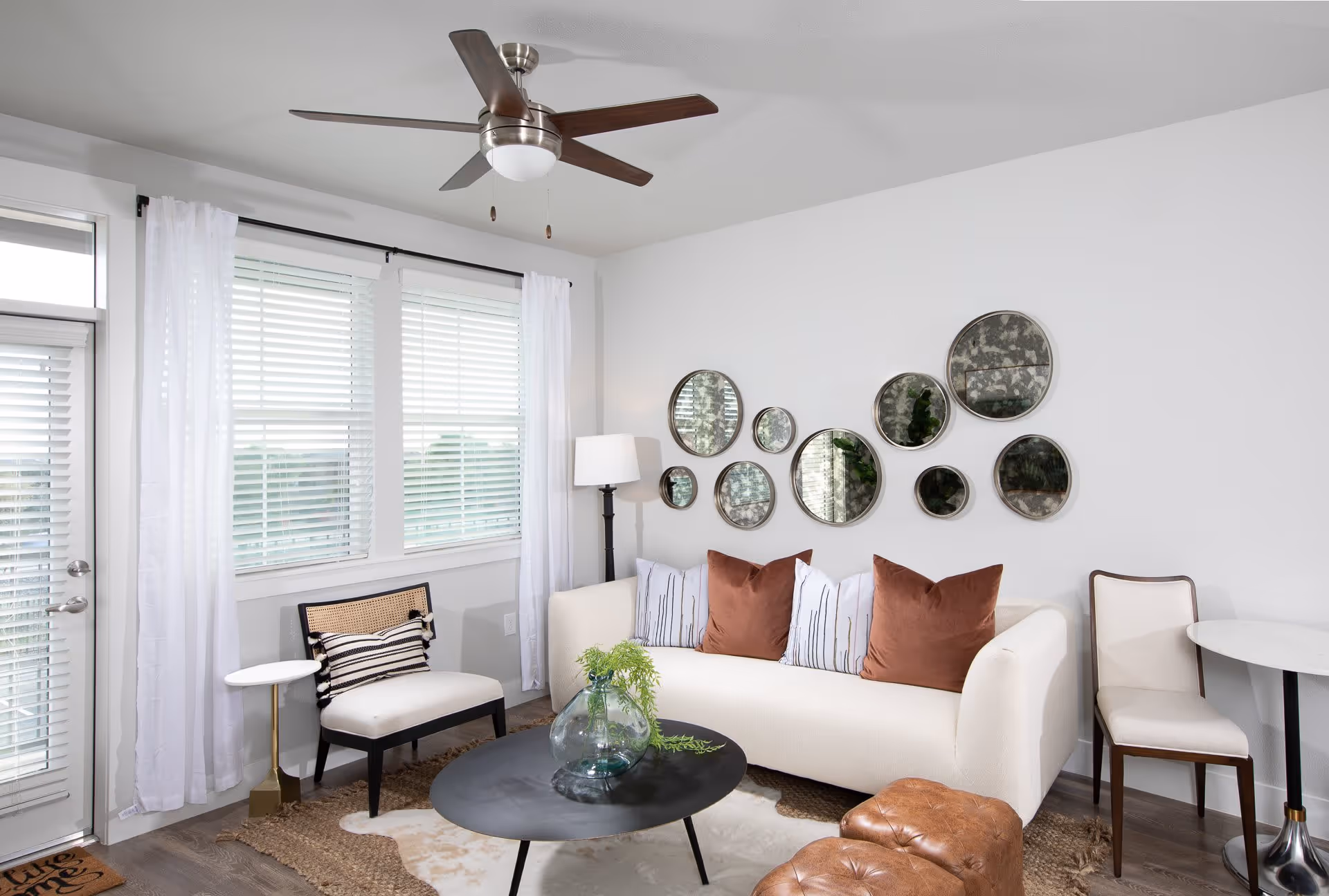 A bright living room with a white sofa adorned with brown and white pillows, a black round coffee table with a glass vase containing greenery, a white cushioned chair with a striped pillow, a floor lamp, and a set of decorative round mirrors on the wall. The room has large windows with white blinds and sheer white curtains, a ceiling fan with wooden blades, and a small round side table with a chair.