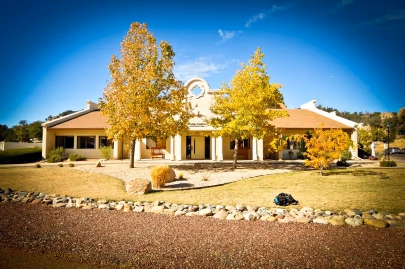Front exterior view of a single-story building with a beige facade and a brown roof, flanked by two trees with yellow autumn leaves. The building has a central entrance with columns and a decorative architectural element above the door. The foreground features a landscaped area with rocks, grass, and a clear blue sky overhead.
