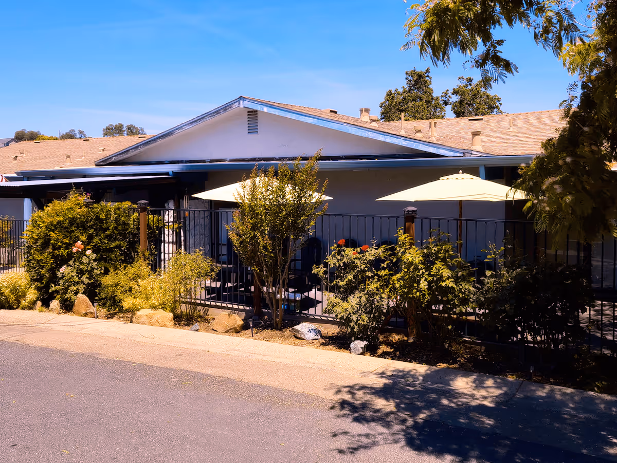 Exterior front of a single-story building with a fenced patio area, umbrellas, and surrounding shrubs and trees.