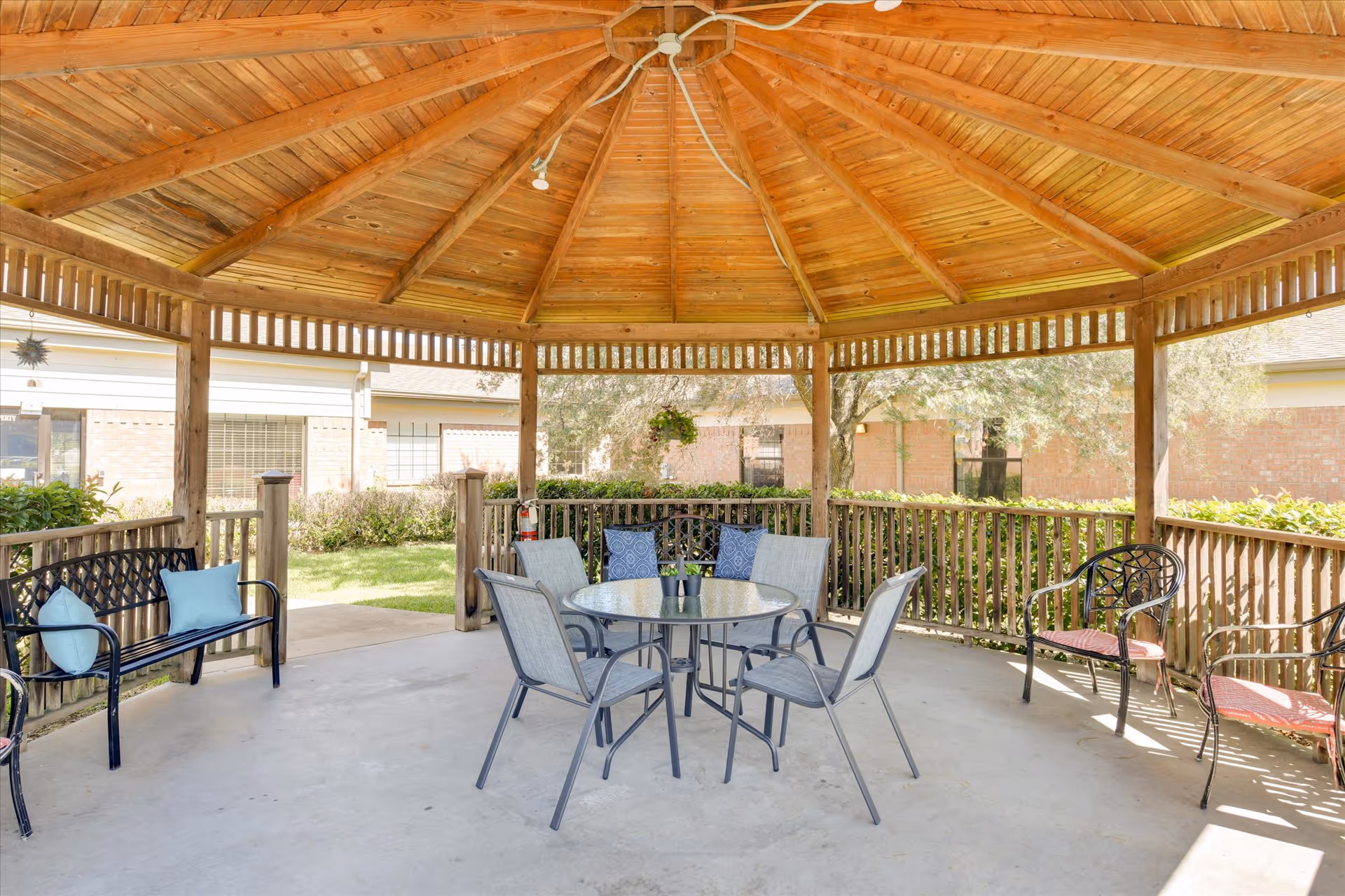 A wooden gazebo with a peaked roof and open sides, furnished with a round glass table surrounded by four chairs, and additional benches and chairs with cushions around the perimeter. The gazebo is situated outdoors with greenery and buildings visible in the background.