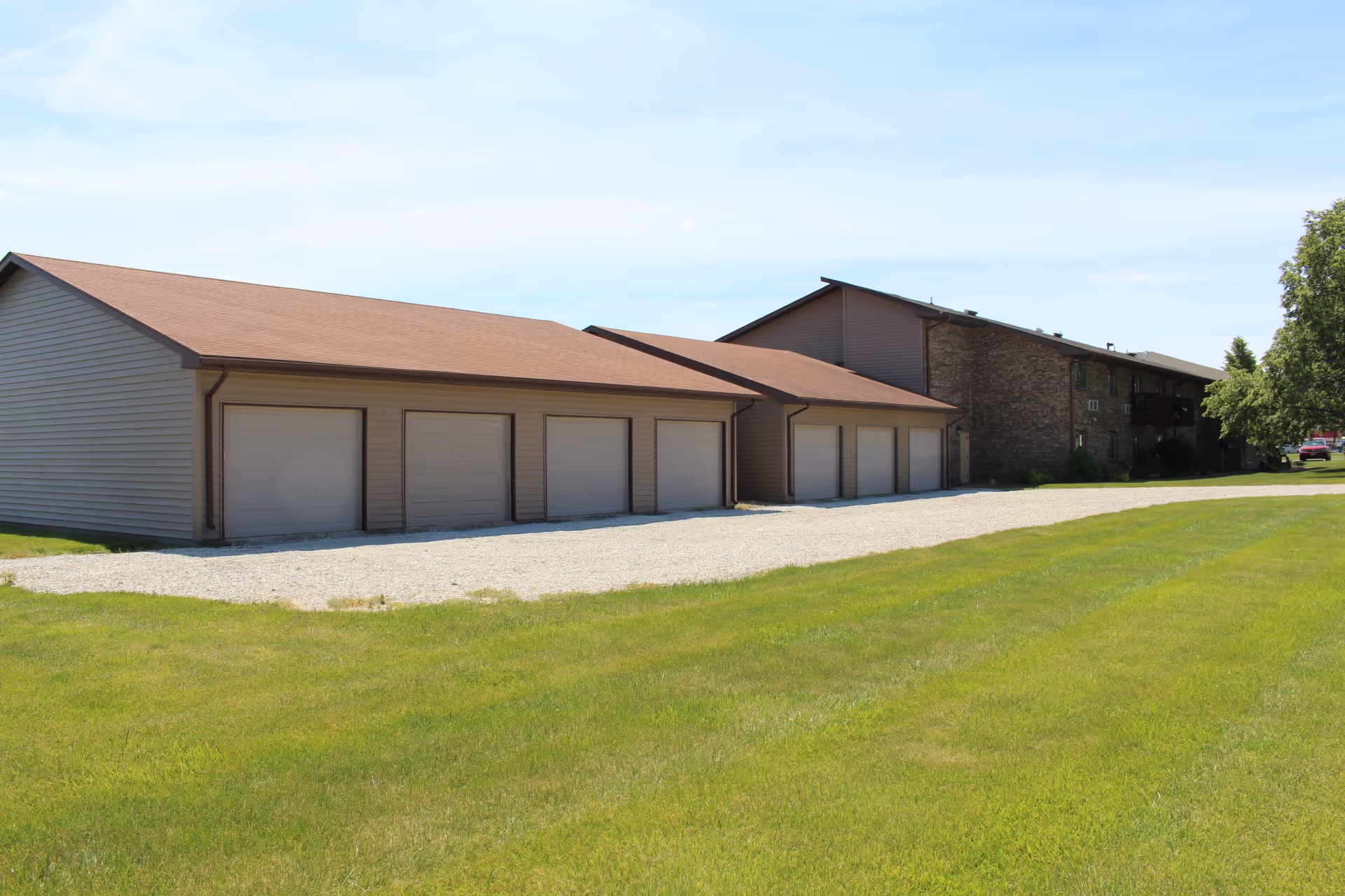 Exterior view of a senior living facility showing a row of closed garage doors attached to a building with a brown roof and beige siding. The building extends into the background with a mix of brick and siding walls. There is a gravel driveway in front of the garages and a well-maintained green lawn in the foreground under a clear blue sky.