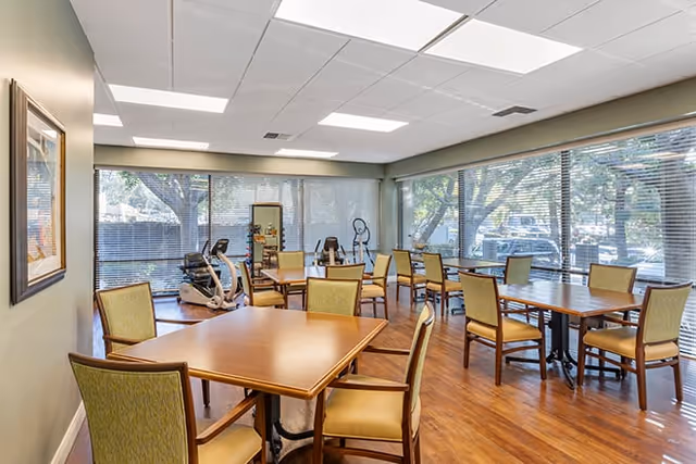 A bright room with large windows on two sides showing trees outside. The room contains several wooden tables with green cushioned chairs arranged around them. In the corner, there is exercise equipment including a stationary bike and an elliptical machine. The floor is wooden, and the ceiling has recessed lighting panels.