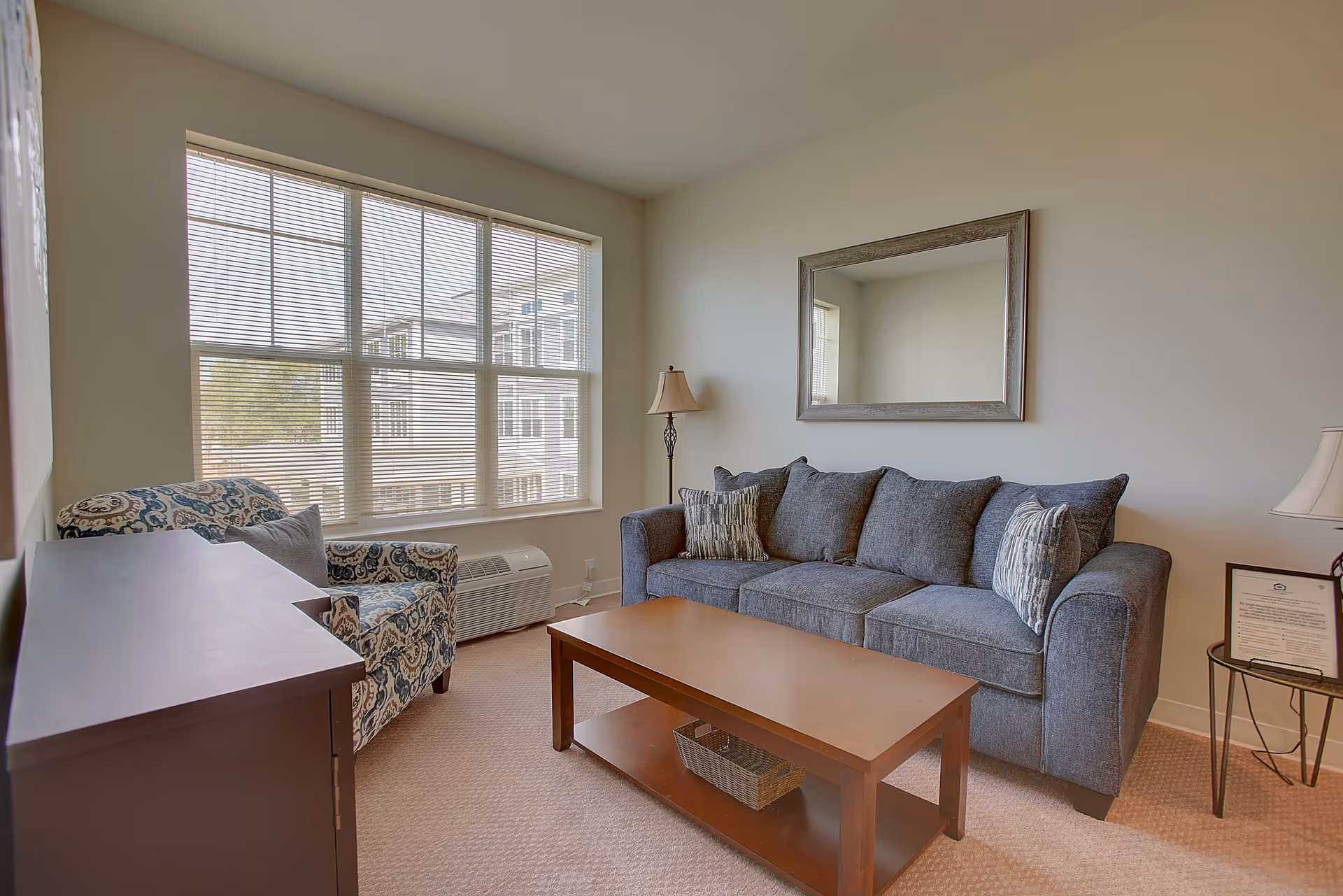 Bright living room with a gray sofa, patterned armchair, wooden coffee table, large window and wall mirror.