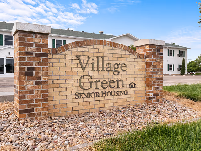 A brick sign for Village Green Senior Housing set in a landscaped area with rocks and grass, with a two-story white building and blue sky in the background.