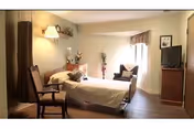 A well-lit bedroom with a single bed, a wooden chair, a small armchair near a window with curtains, a TV on a wooden stand, and decorative items including a wall lamp, framed pictures, and a vase with flowers.