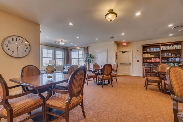 A well-lit common area with several wooden tables and striped cushioned chairs arranged around them. There is a large wall clock showing the time as 11:10, a globe on one of the tables, a bookshelf filled with books, and large windows with curtains letting in natural light.
