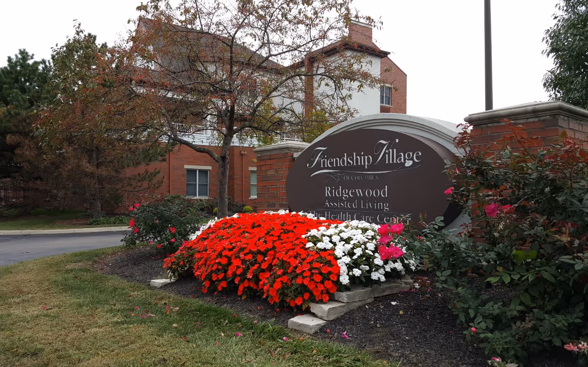 Outdoor view of a senior living facility entrance with a large sign reading 'Friendship Village of Columbus Ridgewood Assisted Living & Health Care Center' surrounded by red, white, and pink flowers and trees, with a brick building in the background.