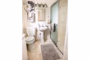 Small bathroom with a toilet, pedestal sink under a mirror, glass-enclosed shower, towel racks, and wall art.