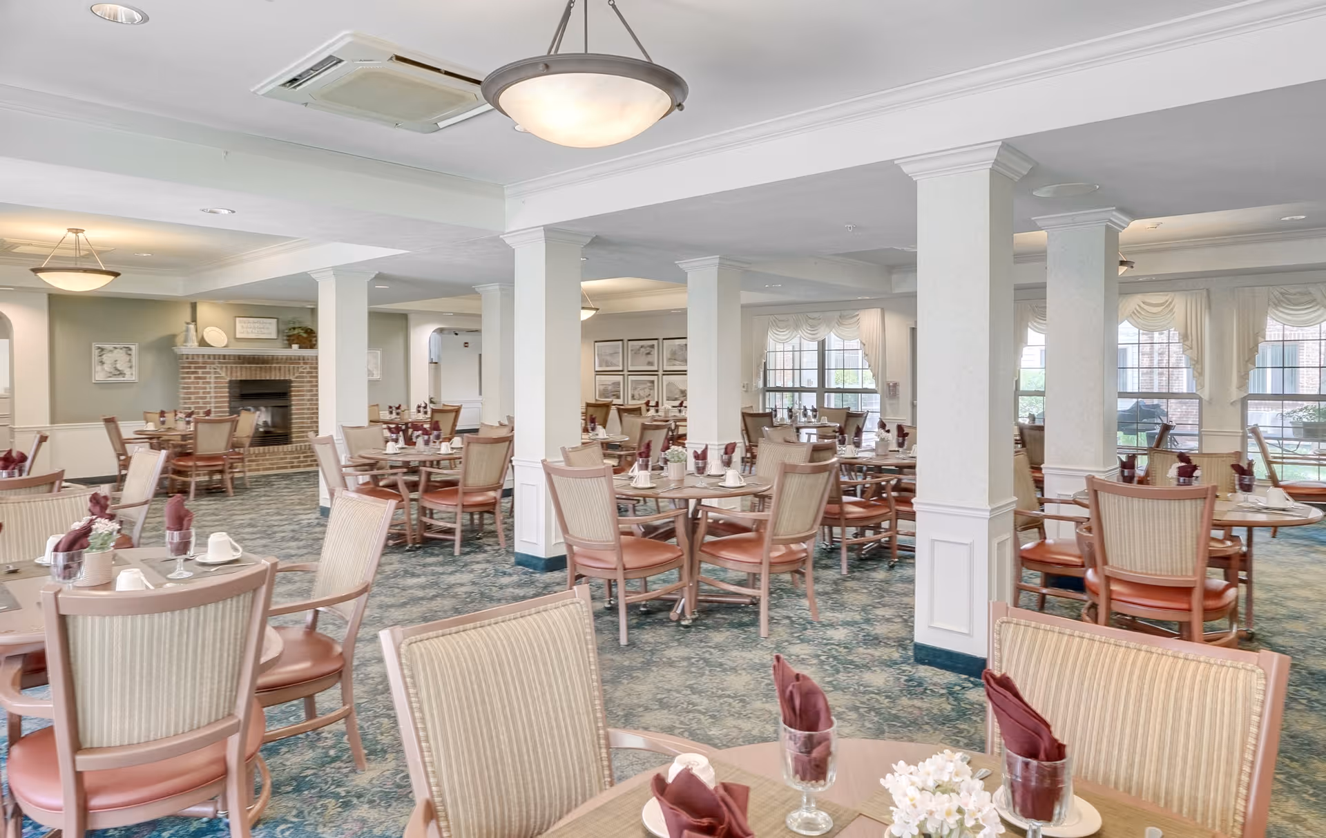 A spacious dining room with multiple round tables and chairs arranged neatly. Each table is set with folded burgundy napkins in glasses, white cups, and small floral centerpieces. The room features white columns, large windows with white curtains, and a brick fireplace in the background. The carpet has a blue and green pattern, and ceiling lights provide soft illumination.