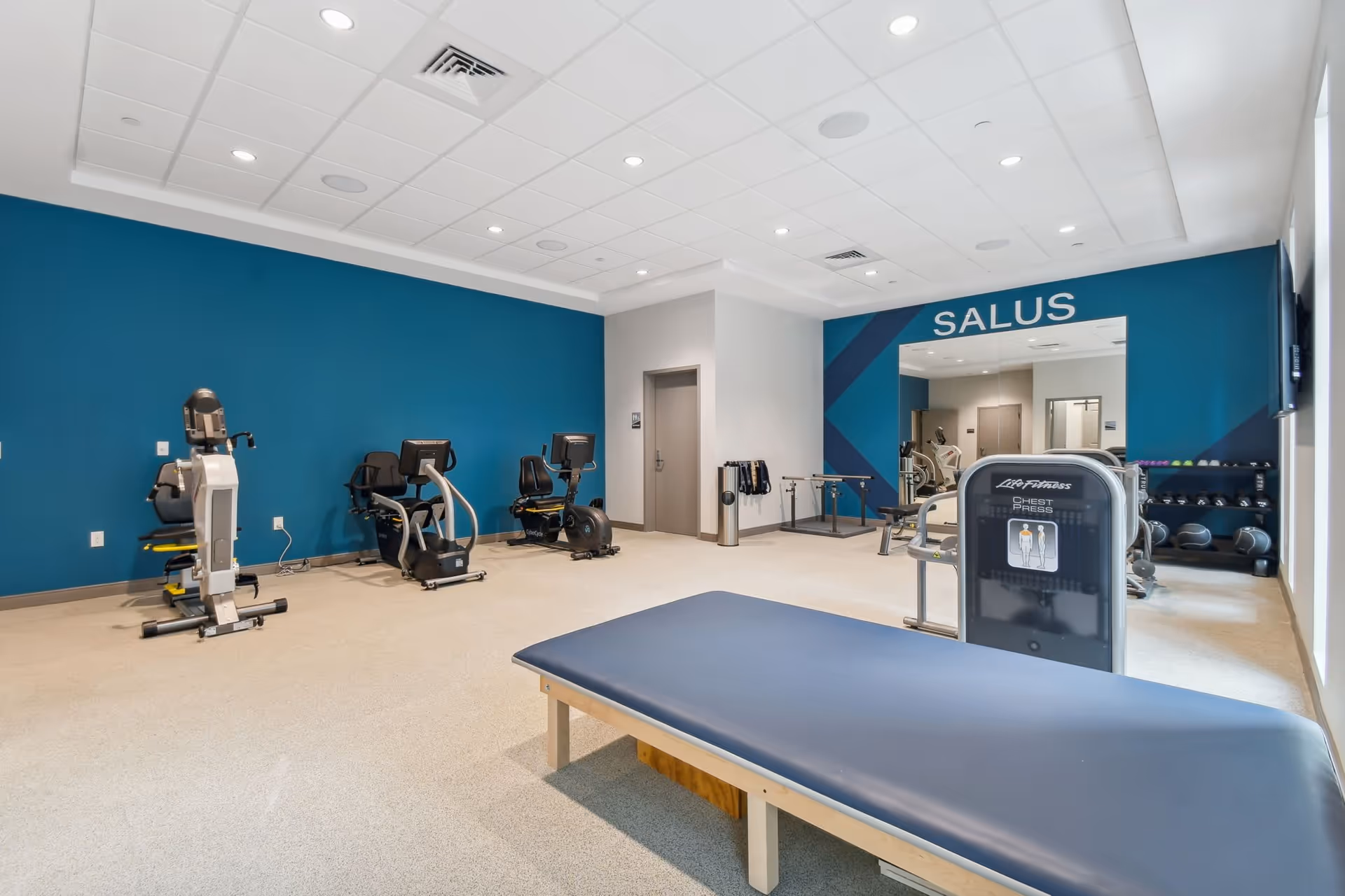 A spacious fitness room with exercise equipment including stationary bikes, a chest press machine, and a padded exercise bench. The walls are painted blue and white, with a large mirror and the word 'SALUS' displayed above it. There are also dumbbells and other fitness accessories visible in the background.