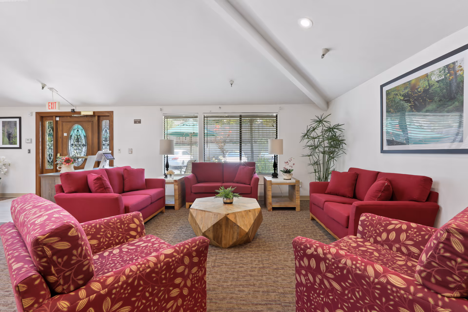 A bright and spacious living room area in Almond Grove Assisted Living featuring red sofas and patterned armchairs arranged around a wooden geometric coffee table. The room has large windows with blinds, two table lamps on wooden side tables, a tall green plant in the corner, and a framed nature photograph on the wall. The entrance door with decorative glass panels is visible in the background.