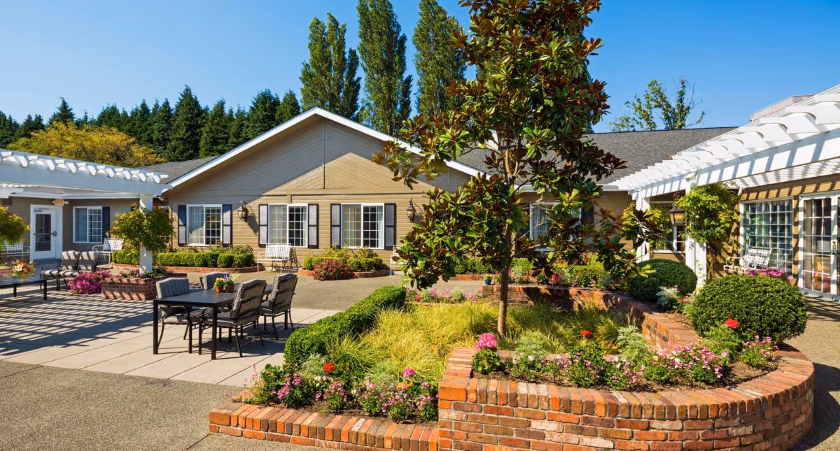 Outdoor courtyard area at Canterbury Gardens Memory Care Community featuring a brick planter with flowers and shrubs, patio tables and chairs, white pergolas, and a beige building with windows and black shutters in the background under a clear blue sky.