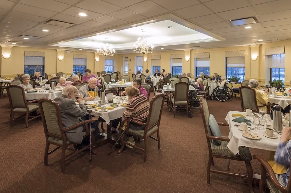 Elderly residents seated at tables eating in a large, well-lit communal dining room.