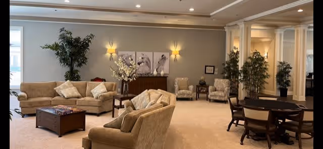 A spacious living room area with beige sofas and armchairs arranged around a wooden coffee table. There are decorative plants, wall sconces, and framed horse artwork on the wall. A round table with chairs is visible on the right side, and the room has a warm, inviting atmosphere with neutral tones and recessed lighting.